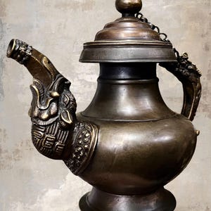 Puede incluir: Tetera antigua de bronce con detalles ornamentales de dragón en el pico y el asa. La tapa está sujeta con una cadena. La tetera tiene un cuerpo bulboso y una base de pedestal. El bronce tiene una pátina oscura y envejecida.