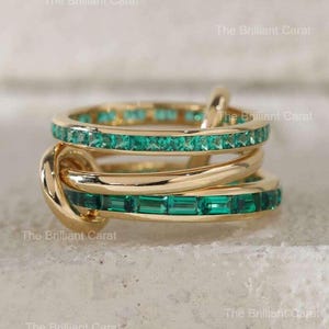 Op de afbeelding: Een goudkleurige ring met drie banden, waarvan twee bezet zijn met smaragdgroene rechthoekige edelstenen. De banden zijn met elkaar verweven, waardoor een uniek knoopontwerp ontstaat. De tekst "The Brilliant Carat" is zichtbaar.
