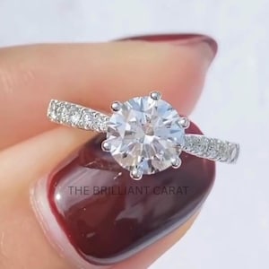 Op de afbeelding: Een close-up van een diamanten verlovingsring. De ring heeft een grote, rond geslepen diamant in een zetting met zes pootjes. De band is van wit metaal en is bezet met kleinere diamanten. De tekst "THE BRILLIANT CARAT" is zichtbaar.