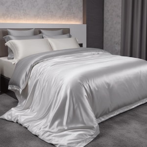 May include: A bed with white and silver satin bedding. The bed has white pillowcases and a silver duvet cover. The bed is in a room with gray walls and a gray carpet.