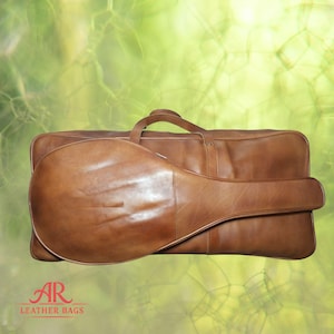 Puede incluir: Un estuche de cuero marrón para instrumentos y una bolsa rectangular a juego. El estuche está diseñado para un instrumento de cuerda, y la bolsa tiene un asa. El logotipo "AR LEATHER BAGS" es visible en la esquina inferior izquierda.