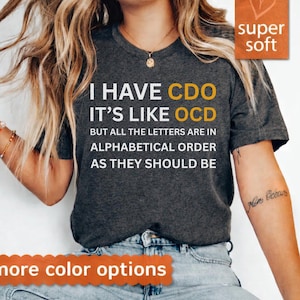 Può includere: T-shirt grigio scuro con la scritta "I HAVE CDO IT'S LIKE OCD BUT ALL THE LETTERS ARE IN ALPHABETICAL ORDER AS THEY SHOULD BE" in bianco e giallo. La maglietta ha un'etichetta "super soft".