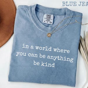 Pode incluir: Uma camiseta azul jeans Comfort Colors com o texto "in a world where you can be anything be kind" em branco. Um colar de ouro com um pingente está sobre a camiseta. Um chapéu marrom e folhas secas estão no fundo.