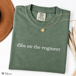 Könnte beinhalten: Ein moosgrünes Kurzarm-T-Shirt mit Rundhalsausschnitt und dem weißen Schriftzug "dibs on the engineer" auf der Brust. Das T-Shirt hat ein Comfort Colors Markenetikett am Innenkragen und besteht aus ringgesponnener Baumwolle.