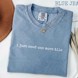 May include: A blue jean coloured short-sleeve t-shirt with a crew neckline. The shirt features the white text "i just need one more tile" printed across the chest. A Comfort Colors brand label is visible at the inner neck, indicating size S.