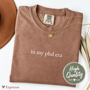以下が含まれることがあります： 胸元にin my phd eraと白で刺繍された茶色の半袖Tシャツ。クルーネックで、襟の内側にはComfort Colorsのブランドラベルが付いています。ゴールドのチェーンネックレスが生地の上に置かれています。