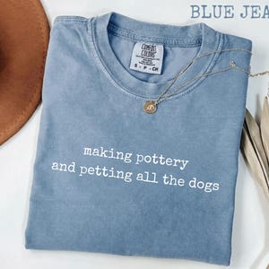 May include: A blue jean colored t-shirt with a crew neckline and the white text making pottery and petting all the dogs printed on the front. The shirt features a Comfort Colors brand label at the inner neck with size S P CH indicated.