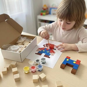 Pode incluir: Uma criança pinta uma imagem pixelizada de um personagem com blocos vermelhos, azuis e castanhos. Potes de tinta de várias cores e blocos de madeira estão sobre a mesa. A imagem está em uma folha de papel branca.