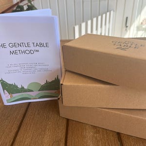 May include: Two brown cardboard boxes are stacked, with the words "THE GENTLE TABLE" printed on the top. A book titled "THE GENTLE TABLE METHOD" is visible, detailing a 90-day nervous system reset guide for recovery.