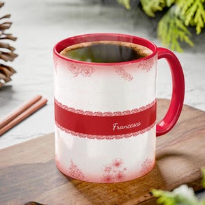 Puede incluir: Una taza de café blanca con asa y borde rojos, llena de café. La taza presenta una banda roja con el nombre "Francesca" y diseños de encaje floral. La taza está sobre una tabla de cortar de madera.