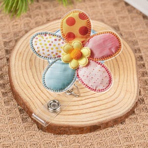 May include: A colorful flower-shaped badge reel with a clear plastic retractable cord. The flower has five petals in various colors and patterns, including yellow with orange polka dots, pink, and blue. The center of the flower is yellow.