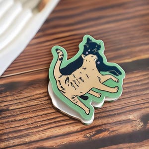 May include: A cat-themed sticker featuring a light brown cat with dark brown markings, overlaid with a dark blue cat silhouette. The sticker has a green border and a white base, set against a wooden surface.