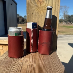 May include: Three maroon leather drink holders. One holds a can, another a can, and the third a brown glass bottle. The holders have white stitching and a fish design. The items are on a wooden surface.