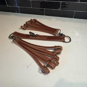 May include: Several brown leather straps with metal hardware, including clasps and rings, are arranged on a white surface. The straps appear to be for a bag or other accessory, with multiple smaller straps attached to a larger one.