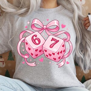 Op de afbeelding: Een grijs gemêleerd sweatshirt met een grafische print van twee roze dobbelstenen met rode hartjespatronen. De dobbelstenen zijn samengebonden met een grote roze strik. Het cijfer zes staat op de linker dobbelsteen en zeven op de rechter.