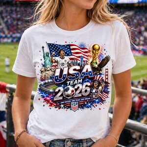 Op de afbeelding: Een wit t-shirt met een grafische print met de tekst USA TEAM 2026 en ONE NATION. ONE TEAM. ONE DREAM. Het ontwerp bevat een voetballer, het Vrijheidsbeeld, Mount Rushmore, een voetbal, een trofee en de Amerikaanse vlag.