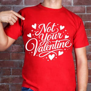 May include: A red t-shirt with the phrase "Not Your Valentine" in white script and heart accents. The shirt is short-sleeved and appears to be made of a soft material. The design is centered on the chest.