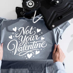 Puede incluir: Una camiseta azul pizarra con texto impreso en blanco que dice Not Your Valentine, rodeado de pequeños iconos de corazones y estrellas. La prenda está colocada bajo una prensa térmica negra, utilizada para aplicar el diseño gráfico.