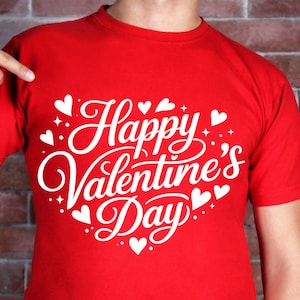 Puede incluir: Una camiseta roja con la frase "Happy Valentine's Day" en blanco, rodeada de corazones blancos y estrellas. La camiseta es de material suave, ideal para el Día de San Valentín.