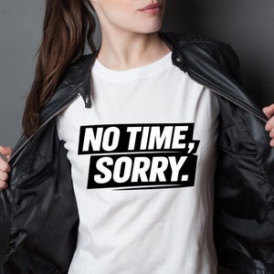 May include: A white t-shirt with the text "NO TIME, SORRY." in a black, bold font. The text is centered within a black rectangle. The shirt is worn under a black leather jacket.