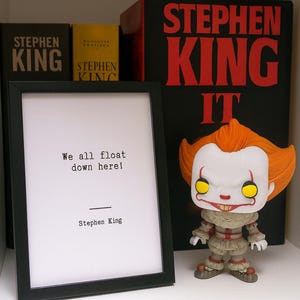 Peut inclure: Une présentation de livres de Stephen King et une citation encadrée : « We all float down here ! » Une figurine de Pennywise est placée à côté des livres. Les livres sont noirs, rouges et jaunes. La citation encadrée est dans un cadre noir.