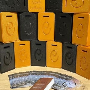 Peut inclure: Plusieurs objets rectangulaires noirs et dorés avec un design en forme de 'C', certains avec une fente en haut. Les objets sont disposés en rangées. Un jeu de cartes avec du texte et un motif est au premier plan.