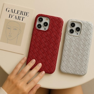 May include: Two phone cases with a woven pattern. One case is a deep red, the other is a light gray. A piece of art with the text "GALERIE D'ART" is in the background.