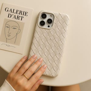 May include: A cream-colored phone case with a woven pattern. The case is next to a card with the text "GALERIE D'ART" and a minimalist line drawing of a face. A gold ring is on a finger.
