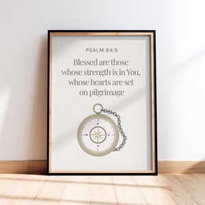 May include: Framed print with the text "PSALM 84:5 Blessed are those whose strength is in You, whose hearts are set on pilgrimage." A compass illustration is below the text, with a chain attached. The frame is black.