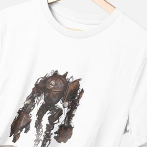 May include: A white short-sleeved t-shirt featuring a graphic print of a weathered, brown mechanical robot. The robot has a rounded torso with a skull emblem, large claw-like hands, and visible wiring details. The shirt is displayed on a hanger.
