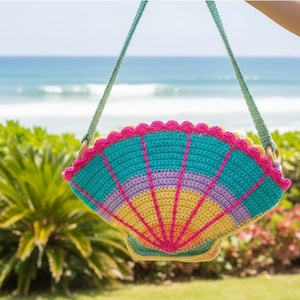 May include: A colorful, crocheted seashell-shaped bag with a green strap. The bag features a scalloped edge and a fan-like design in shades of teal, pink, purple, and yellow. The background shows a beach scene with green foliage and the ocean.