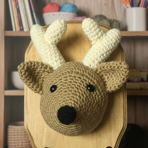 May include: A handcrafted, crochet deer head mounted on a wooden plaque. The deer head is brown with cream-colored antlers and a black nose and eyes. The plaque is supported by a black stand, creating a unique wall decor piece.