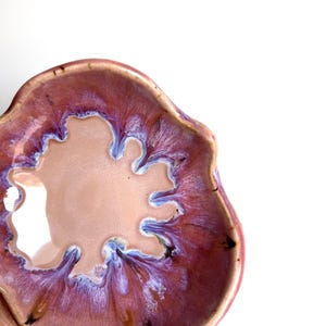 Pode incluir: Uma tigela de cerâmica com formato orgânico irregular, apresentando um centro liso bege-claro que transita para uma borda brilhante e vibrante de magenta profundo, roxo e estrias de esmalte branco que irradiam para a borda ondulada e texturizada.