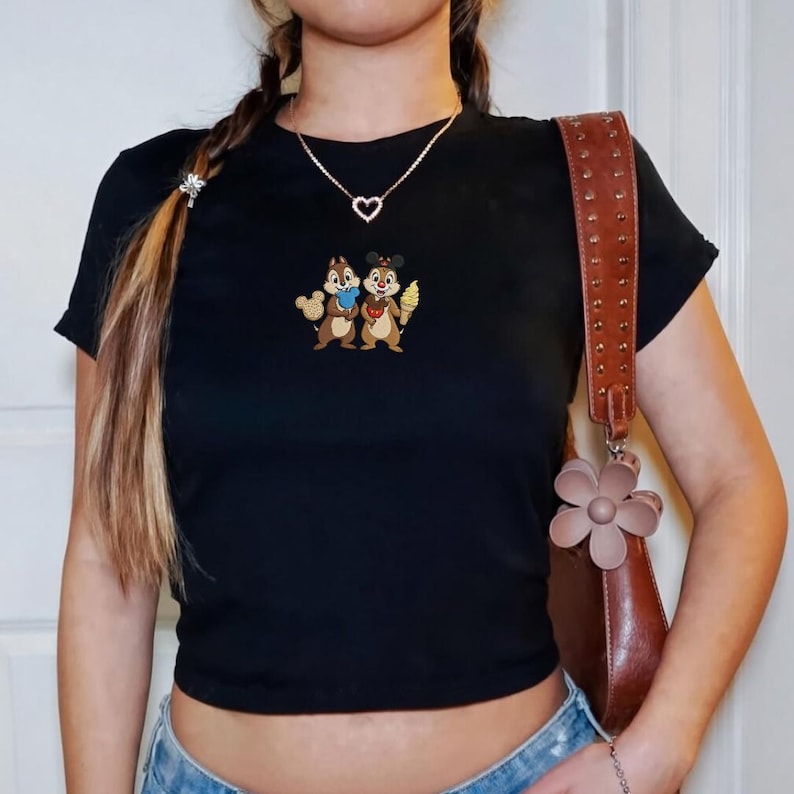 May include: Black crop top featuring an embroidered design of two cartoon chipmunks, one holding a cookie and the other an ice cream cone. A gold heart-shaped necklace and a brown shoulder bag with a flower charm are also visible.