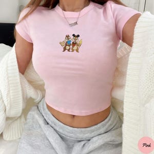 May include: A light pink crop top featuring a cartoon graphic of two chipmunks holding ice cream and a heart. The shirt has short sleeves and a crew neck. The word "Pink" is in a pink circle in the bottom right corner.