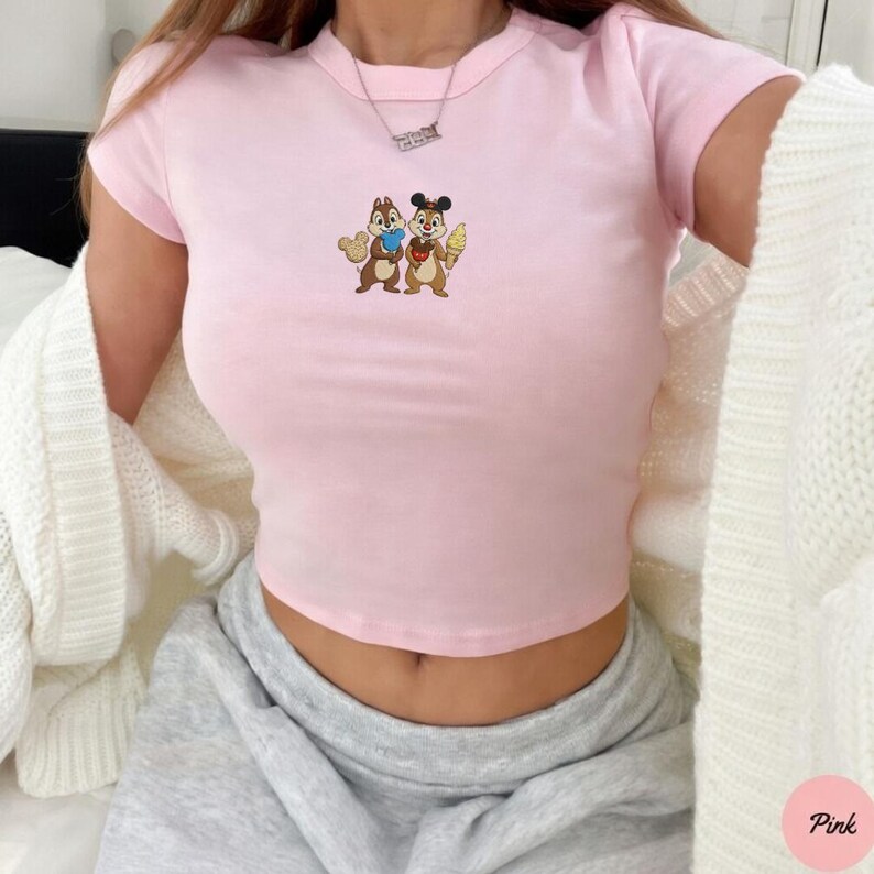 May include: A light pink crop top featuring a cartoon graphic of two chipmunks holding ice cream and a heart. The shirt has short sleeves and a crew neck. The word "Pink" is in a pink circle in the bottom right corner.
