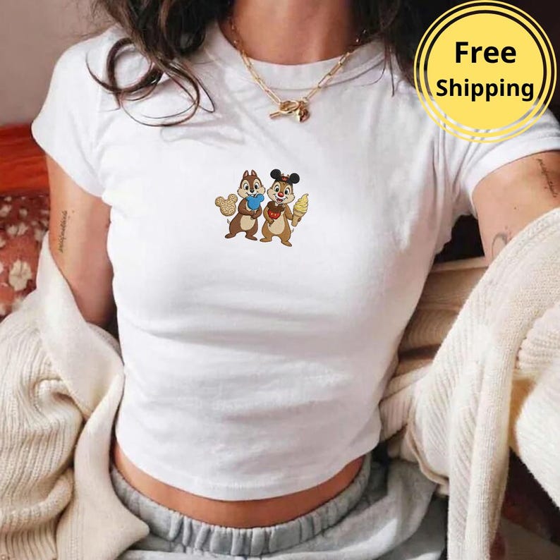 May include: White crop top featuring a colorful embroidered design of two cartoon chipmunks. One chipmunk holds a blue object, the other an ice cream cone. The shirt has a crew neck and short sleeves.
