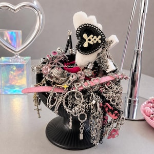 Könnte beinhalten: Eine schwarze Fußschale gefüllt mit einer Sammlung silberfarbener Ketten, rosa Lederriemen und Anhängern. Zu den Details gehören ein weißes, flauschiges Herzornament mit schwarzem Herzmittelstück, rosa Strasssteinen und einem kleinen silbernen Kreuz.