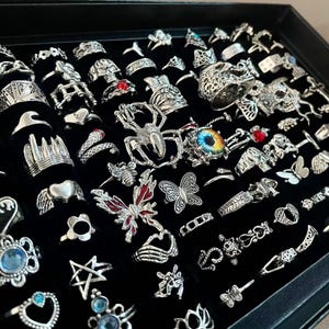 Puede incluir: Una colección de anillos plateados exhibidos en una bandeja negra. Los anillos presentan varios diseños, incluyendo calaveras, arañas y mariposas. Algunos anillos tienen detalles coloridos, como piedras rojas y azules.