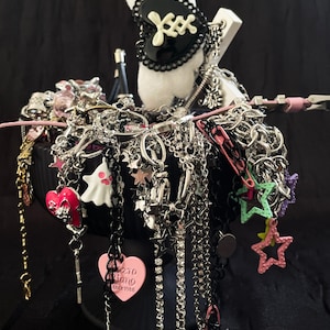Puede incluir: Una colección de collares y pulseras plateadas con varios dijes, incluyendo corazones, estrellas y un fantasma. Un cuenco negro contiene las joyas, con un artículo de felpa blanco y un accesorio en blanco y negro en la parte superior. Se ven acentos rosas y negros.