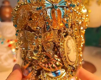 May include: A decorative jar overflowing with gold-toned embellishments. It features butterfly and floral motifs, along with blue and clear gemstones. A red flower adorns the top. The jar is held by hands.
