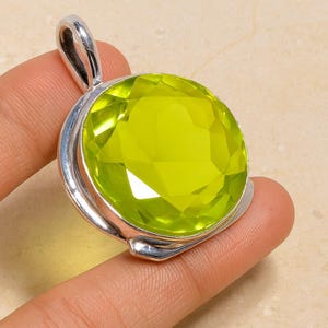 May include: A round, faceted, lime-green gemstone pendant with a silver-coloured bail. The gemstone is set in a silver bezel. The pendant is held by a person's fingers against a neutral background. Jewellery.