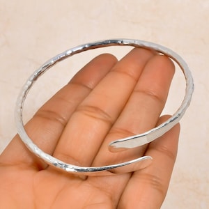 Könnte beinhalten: Ein silberfarbenes Armreif-Armband mit gehämmertem Muster. Das offene Design hat eine leicht ovale Form und eine strukturierte Oberfläche. Das Armband wird vor einem hellen Hintergrund gezeigt.