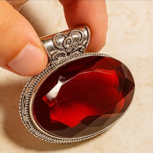 Könnte beinhalten: Ein silberner Anhänger mit einem ovalen, facettierten, tiefroten Edelstein. Der Anhänger hat eine verzierte Öse und einen detaillierten silbernen Rand. Der Edelstein ist in Silber gefasst. Der Anhänger wird von einer Person gehalten.