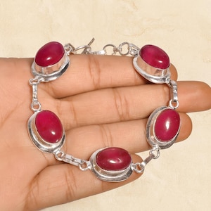 Puede incluir: Una pulsera de plata con piedras preciosas ovaladas de color rojo intenso. Las piedras están engastadas en biseles individuales de plata y conectadas por eslabones de plata. La pulsera se muestra sobre un fondo claro.