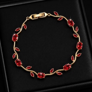 Könnte beinhalten: Ein goldfarbenes Armband mit einer rankenartigen Struktur, kleinen spitzen Blättern und ovalen, leuchtend roten Edelsteinen, die entlang der Kette eingefasst sind. Das Schmuckstück verfügt über einen sicheren Metallverschluss.