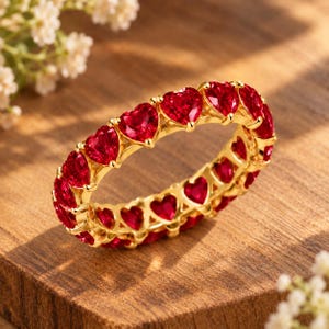 Può includere: Una fede in oro con una fila continua di pietre preziose rosse a forma di cuore. L'anello è realizzato con una montatura in metallo che tiene in posizione ogni singola pietra tagliata a cuore, creando un design circolare di colore rosso.