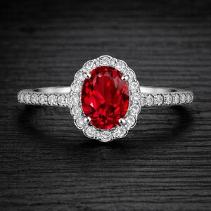 Peut inclure: Une bague en argent ornée d'une grande pierre rouge vif de forme ovale, entourée d'un halo de petites pierres claires. La monture est également sertie de petites pierres claires, créant un effet scintillant. Le fond est sombre.