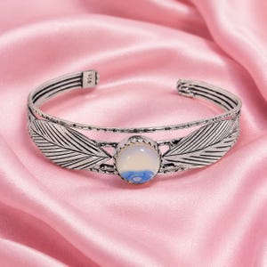 Puede incluir: Una pulsera de brazalete de plata con una piedra cabujón redonda central con un patrón de remolinos blancos lechosos y azules. La banda está decorada con motivos de hojas grabados en ambos lados e incluye una pequeña etiqueta con el 925.