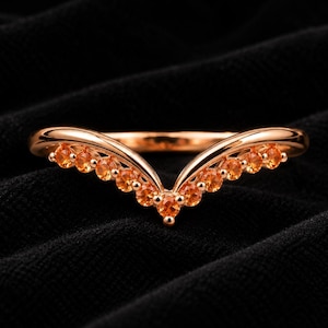 Op de afbeelding: Een V-vormige ring van roségoud met een reeks kleine, ronde, oranje edelstenen die langs de chevron-stijl band zijn gezet. Het metaal heeft een gepolijste, reflecterende afwerking en de stenen zijn symmetrisch gerangschikt langs het ontwerp.